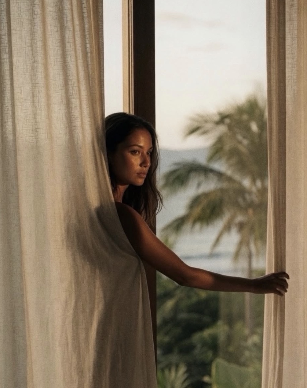 A guest drawing back sheer linen curtains to the ocean and palms beyond